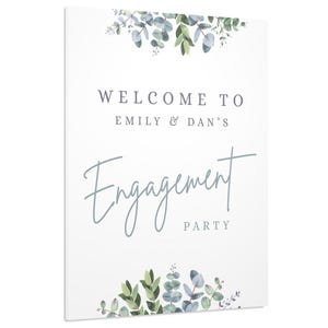 May include: A white engagement party sign with the text "Welcome to Emily & Dan's Engagement Party" in a script font. The sign is decorated with green and blue watercolor eucalyptus leaves.