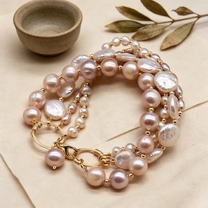 Puede incluir: Una pulsera de múltiples hilos con perlas rosas y blancas de varios tamaños, ensartadas con alambre y detalles dorados. La pulsera tiene un cierre dorado y se exhibe sobre una superficie neutra.