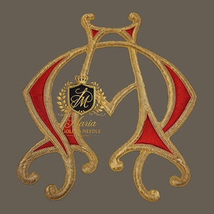 Op de afbeelding: Een sierlijk, goudkleurig monogram met rode accenten, met een gestileerd "AM"-ontwerp. Het monogram is geborduurd met ingewikkelde details en bevat een wapen met een kroon en de tekst "Maria GOLDEN NEEDLE".