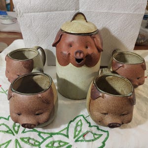 Puede incluir: Un juego de cocina de cerámica con temática de cerdo que incluye un tarro grande con tapa y cuatro tazas a juego. El juego tiene una combinación de colores marrón y beige, con la cara y las orejas del cerdo en cada pieza.