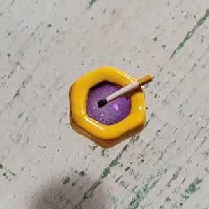 May include: A small yellow hexagonal ashtray with a purple interior and a partially smoked cigarette resting inside.