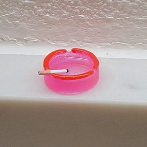May include: A round, translucent pink ashtray with a bright orange rim. The ashtray features a single notch on the edge, which holds a small, white cigarette with a tan filter and a dark, burnt tip resting horizontally across the opening.