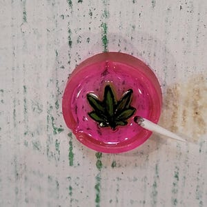 May include: A circular, translucent pink resin ashtray featuring a dark green cannabis leaf graphic embedded in the center. A small, white, cigarette-shaped object rests on the rim of the ashtray, pointing toward the center leaf design.