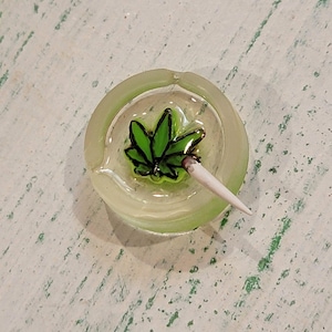 May include: A round, translucent light green ashtray featuring a central green cannabis leaf graphic with black outlines. A small, white, pointed object resembling a cigarette is resting on the edge of the ashtray, pointing toward the center.