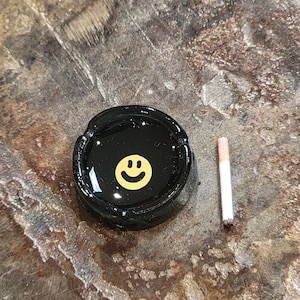 May include: Black ashtray with a yellow smiley face design. A white cigarette is next to the ashtray.