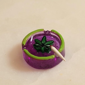 May include: A round, translucent purple ashtray featuring a bright green rim and a central green leaf-shaped emblem. A small, white, cone-shaped object rests on the edge of the ashtray, extending toward the center of the purple basin.