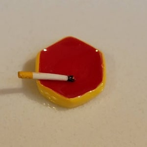May include: A small, yellow hexagonal ashtray with a red interior. A white cigarette is resting in the ashtray.