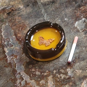 May include: A small, round, yellow ashtray with a black rim. A small, orange butterfly is embedded in the center of the ashtray. A white cigarette with a red tip is lying on a brown surface next to the ashtray.