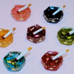 May include: Six small hexagonal ashtrays with a cigarette resting in each. The ashtrays are made of glass and have a sparkly finish. The colors of the ashtrays are pink, black, orange, maroon, green, and red.
