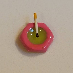 May include: A pink ceramic ashtray with a yellow and black cigarette inside.