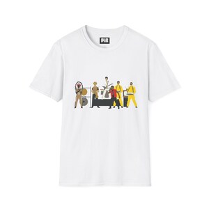 以下が含まれることがあります： ステージ上の音楽バンドのグラフィックイラストが描かれた白い半袖Tシャツ。バンドメンバーは、黄色いジャンプスーツや羽飾りのついたヘッドドレスなど様々な衣装を身にまとい、楽器や機材が一緒に描かれています。