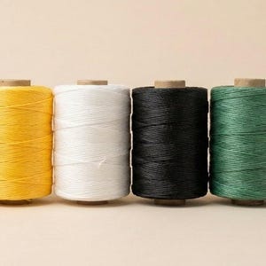 May include: Six spools of colorful thread in red, yellow, white, black, green, and blue. Each spool is cylindrical, with the thread neatly wound around a central core. The threads are tightly packed, and the colors are vibrant against a neutral background.