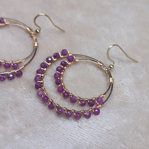 May include: Gold-tone hoop earrings with a double-circle design. The hoops are adorned with small, faceted, purple gemstones. The earrings have a hook closure and are displayed on a soft, white surface. These earrings are a stylish accessory.