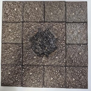 Op de afbeelding: Opstelling van zestien vierkante tegels met een getextureerd, gespikkeld oppervlak in grijze en bruine tinten. De centrale tegel heeft een donker, getextureerd ontwerp dat lijkt op een verkoolde of verbrande zone. De tegels zijn in een rasterpatroon gerangschikt.