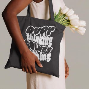 May include: A black tote bag with the text "Overthinking Solves Nothing" in white. The bag is held by a person and contains a bouquet of white tulips. The tote bag is made of a sturdy material.