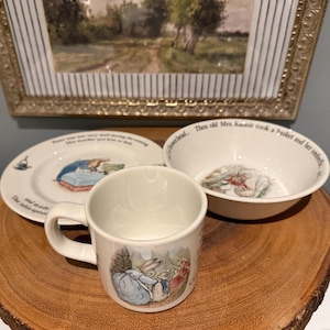 Puede incluir: Un juego de taza, plato y cuenco de cerámica blanca con ilustraciones de personajes de Peter Rabbit. El juego incluye una taza pequeña con asa, un plato y un cuenco. Las ilustraciones son en colores suaves.
