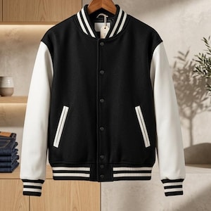 May include: A black varsity jacket with white sleeves, featuring a snap-button front closure, two side pockets with white trim, and a ribbed collar, cuffs, and hem with white stripes. The jacket is displayed on a wooden hanger against a neutral background.