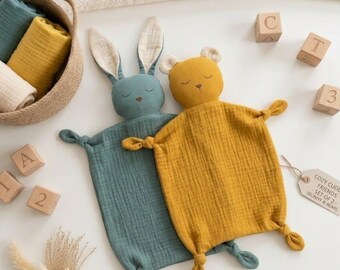 May include: A teal bunny and a mustard yellow bear security blanket, both made of textured fabric with knotted corners. A tag attached to the bear reads COZY CUDDLE FRIENDS - SET OF 2 - (BUNNY & BEAR). Nearby are wooden blocks with letters and numbers.