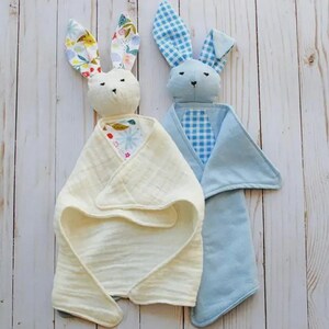 May include: Two plush security blankets shaped like rabbits. One rabbit is cream-coloured with a floral-patterned inner ear and a textured muslin blanket body. The second rabbit is light blue with a blue gingham inner ear and a solid blue blanket.