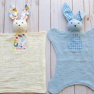 May include: Two baby security blankets, each with a plush bunny head. One is cream-colored with floral ears and a floral patch. The other is light blue with gingham ears and a gingham patch. Both have a soft, textured appearance.
