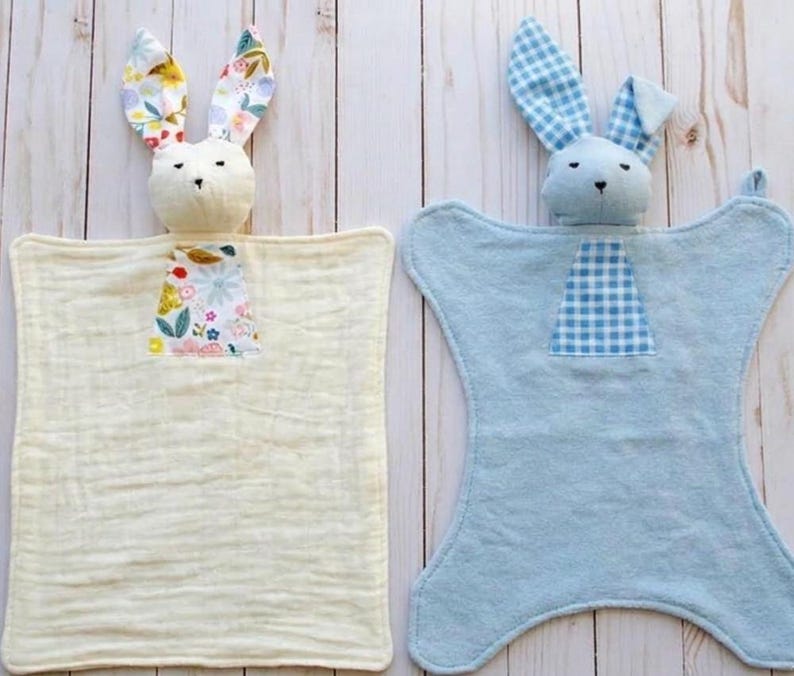 May include: Two baby security blankets, each with a plush bunny head. One is cream-colored with floral ears and a floral patch. The other is light blue with gingham ears and a gingham patch. Both have a soft, textured appearance.