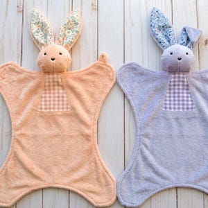 May include: Two plush bunny security blankets. One is peach with floral print ears and a gingham patch. The other is lavender with floral print ears and a gingham patch. Both have a soft, textured appearance.