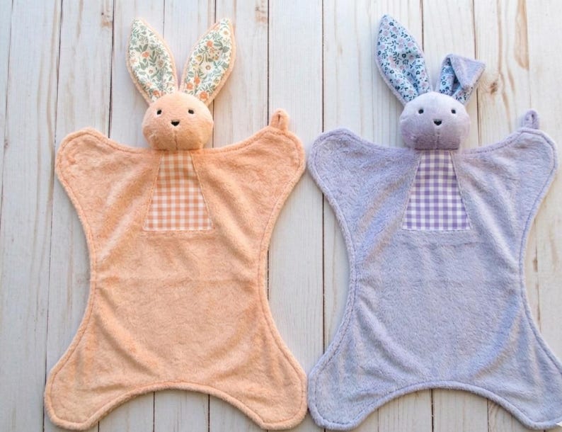 May include: Two plush bunny security blankets. One is peach with floral print ears and a gingham patch. The other is lavender with floral print ears and a gingham patch. Both have a soft, textured appearance.