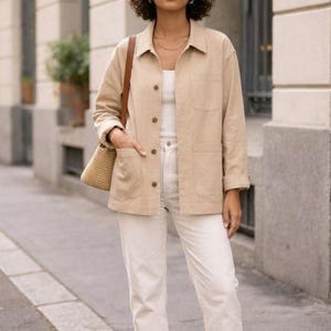 May include: A woman in a tan jacket, white top, and white jeans, with a small straw bag and a brown leather shoulder bag. The jacket has large pockets and button closures. The outfit is casual and stylish, suitable for a day out.