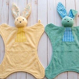 May include: Two plush bunny security blankets. One is yellow with floral print ears and a yellow gingham patch. The other is teal with blue gingham ears and a blue gingham patch. Both have a bunny head and a soft, textured body.