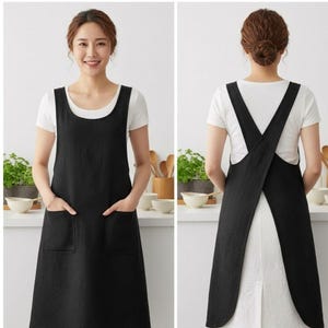Puede incluir: Un delantal negro con escote redondo y dos bolsillos frontales de parche. El diseño incluye tirantes anchos que se cruzan en la espalda, creando un estilo abierto. El delantal se lleva sobre una camiseta blanca de manga corta.