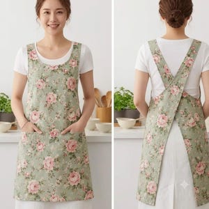 May include: A sage green apron featuring a repeating pattern of pink roses and green leaves. The apron has a scoop neckline, two front patch pockets, and wide shoulder straps that cross over the back in a pinafore style, worn over a white t-shirt.
