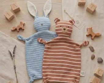 May include: Two soft, flat plush toys shaped like a rabbit and a bear. The rabbit is blue with white horizontal stripes, and the bear is brown with white horizontal stripes. Both have knotted corners, embroidered facial features, and a flat body.