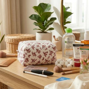 May include: A floral print cosmetic bag with a zipper closure sits on a wooden surface, alongside makeup brushes, a comb, and skincare products. A bottle of Garnier Micellar Water is visible. A potted plant and a mirror are in the background.