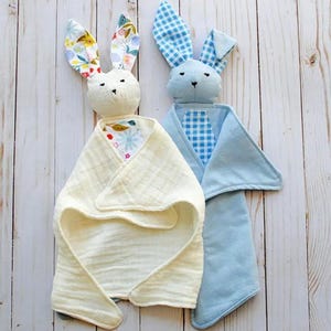 May include: Two handmade bunny security blankets. One bunny is cream-colored with floral print ears and a cream-colored blanket. The other is blue with gingham ears and a blue blanket. The blankets are on a light wood surface.