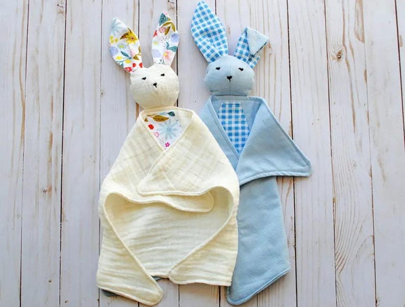 May include: Two handmade bunny security blankets. One bunny is cream-colored with floral print ears and a cream-colored blanket. The other is blue with gingham ears and a blue blanket. The blankets are on a light wood surface.