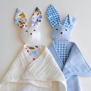 May include: Two plush bunny security blankets. One bunny is white with floral print ears and a white blanket. The other is blue with gingham ears and a blue blanket. Both have embroidered faces.