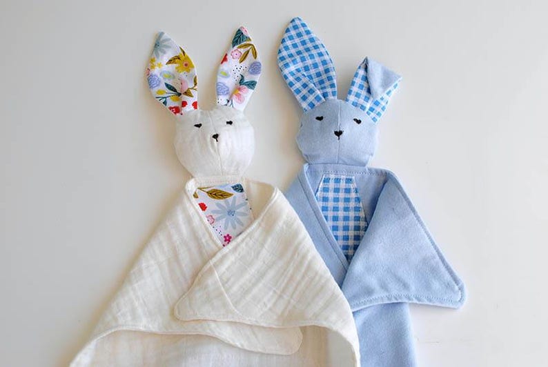 May include: Two plush bunny security blankets. One bunny is white with floral print ears and a white blanket. The other is blue with gingham ears and a blue blanket. Both have embroidered faces.