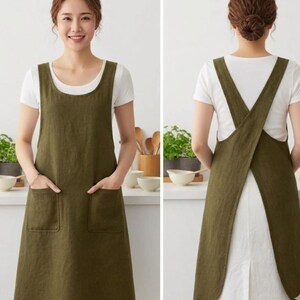 Puede incluir: Un delantal de color verde oliva con escote redondo y dos grandes bolsillos frontales. El diseño incluye tirantes anchos que se cruzan en la espalda, creando un estilo de espalda abierta que cubre completamente la parte frontal del torso.