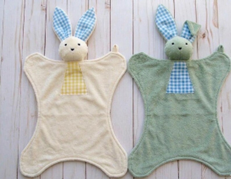 May include: Two baby security blankets, each featuring a plush bunny head and ears with a gingham pattern. One blanket is cream-colored with yellow gingham, the other is sage green with blue gingham. The blankets have a unique shape.