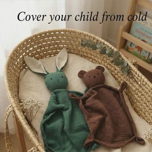 May include: Two soft security blankets shaped like animals, one dark green rabbit with long ears and one brown bear, both featuring embroidered facial details and crinkled fabric textures. The text "Cover your child from cold" is visible above.