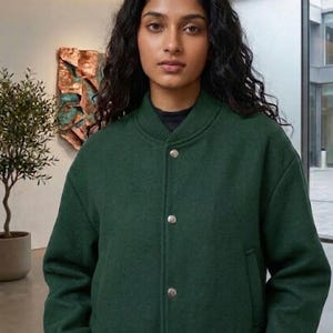 May include: A dark green bomber jacket with a ribbed collar and cuffs. The jacket has a button-up front with silver buttons. The person in the image is wearing a black shirt underneath the jacket.