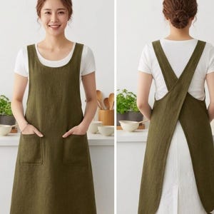 May include: Olive green apron with two front pockets and a cross-back design. The apron is made of a textured fabric and is worn over a white shirt. The apron is designed for kitchen or craft use.