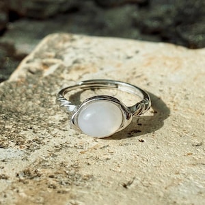 May include: A silver ring featuring a white, oval gemstone. The band has a twisted pattern, and the stone is secured by a fine setting. The ring is shown on a textured, light-coloured surface. The ring is suitable for everyday wear.