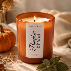 Puede incluir: Una vela encendida en un frasco de vidrio naranja translúcido con una etiqueta blanca que dice Taryn Philip Pumpkin & Patchouli Hand Poured In Oregon 7.5oz/213g. La vela tiene una mecha encendida y está llena de cera de color naranja.
