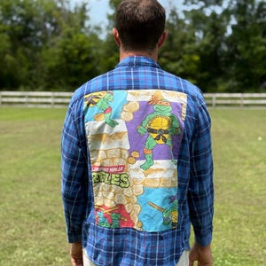 Pode incluir: Uma camisa de manga comprida xadrez azul e preta com um grande painel gráfico retangular nas costas. O gráfico exibe personagens das Tartarugas Ninja, texturas de parede de pedra e o texto Teenage Mutant Ninja Turtles.