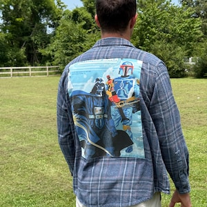 May include: A blue and grey plaid long-sleeve button-down shirt featuring a large rectangular graphic on the back. The graphic displays Star Wars characters Darth Vader and Boba Fett in blue, black, and orange tones with the Star Wars logo text.