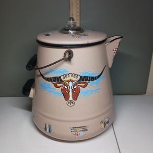 May include: A beige enamel coffee pot with a black rim, a clear glass knob on the lid, and a black handle. The pot features a large steer skull graphic in red, white, and black against a blue brushstroke, with small kitchen utensil icons below.
