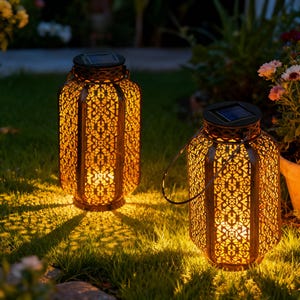 May include: Two bronze-colored solar lanterns with intricate cut-out patterns, casting warm light on the surrounding green grass. Each lantern has a handle and a solar panel on top. The lanterns are placed outdoors near plants.