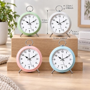 May include: Four round analog quartz alarm clocks in pastel green, white, pink, and light blue. Each clock features a white face with black numerals, a silver ring handle, and two silver feet. Dimensions are 5.31 inches high and 4.84 inches wide.