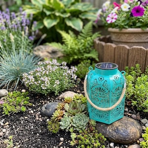 May include: A hexagonal turquoise metal lantern featuring intricate laser-cut floral patterns, a solar panel on the top lid, and a thick beige rope handle, resting on dark soil among various green succulents and garden plants.